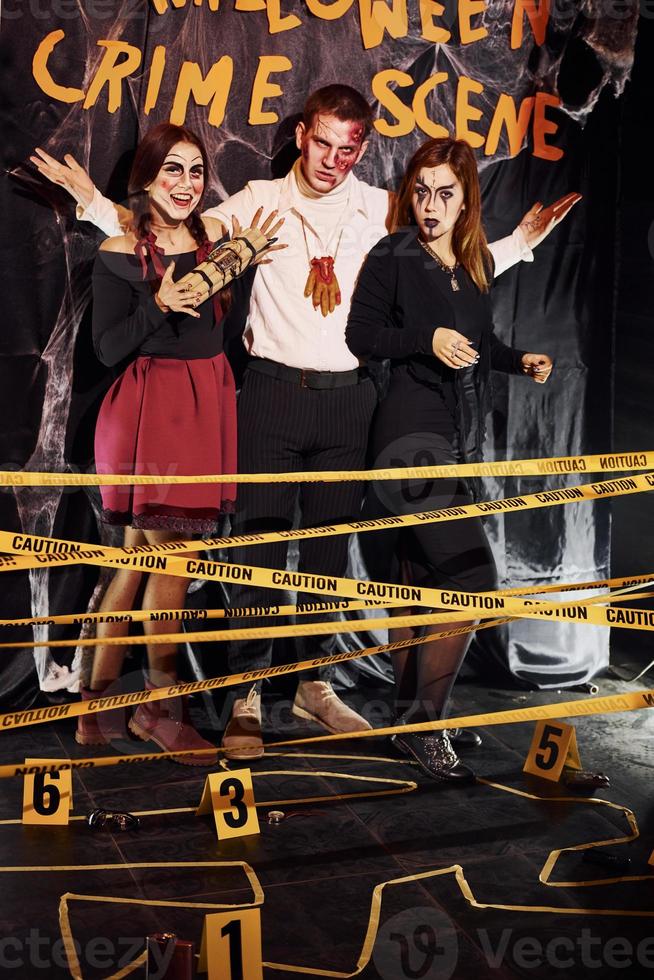 Standing against crime scene wall. Friends is on the thematic halloween party in scary makeup and costumes photo