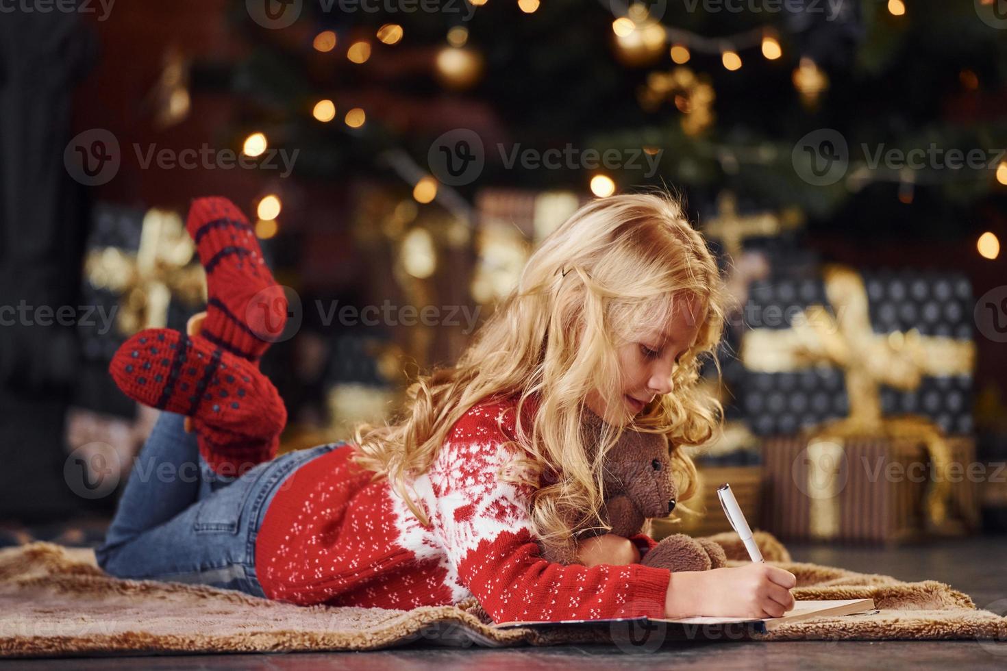 Cute little girl in red festive sweater lying down with teddy bear indoors celebrating new year photo