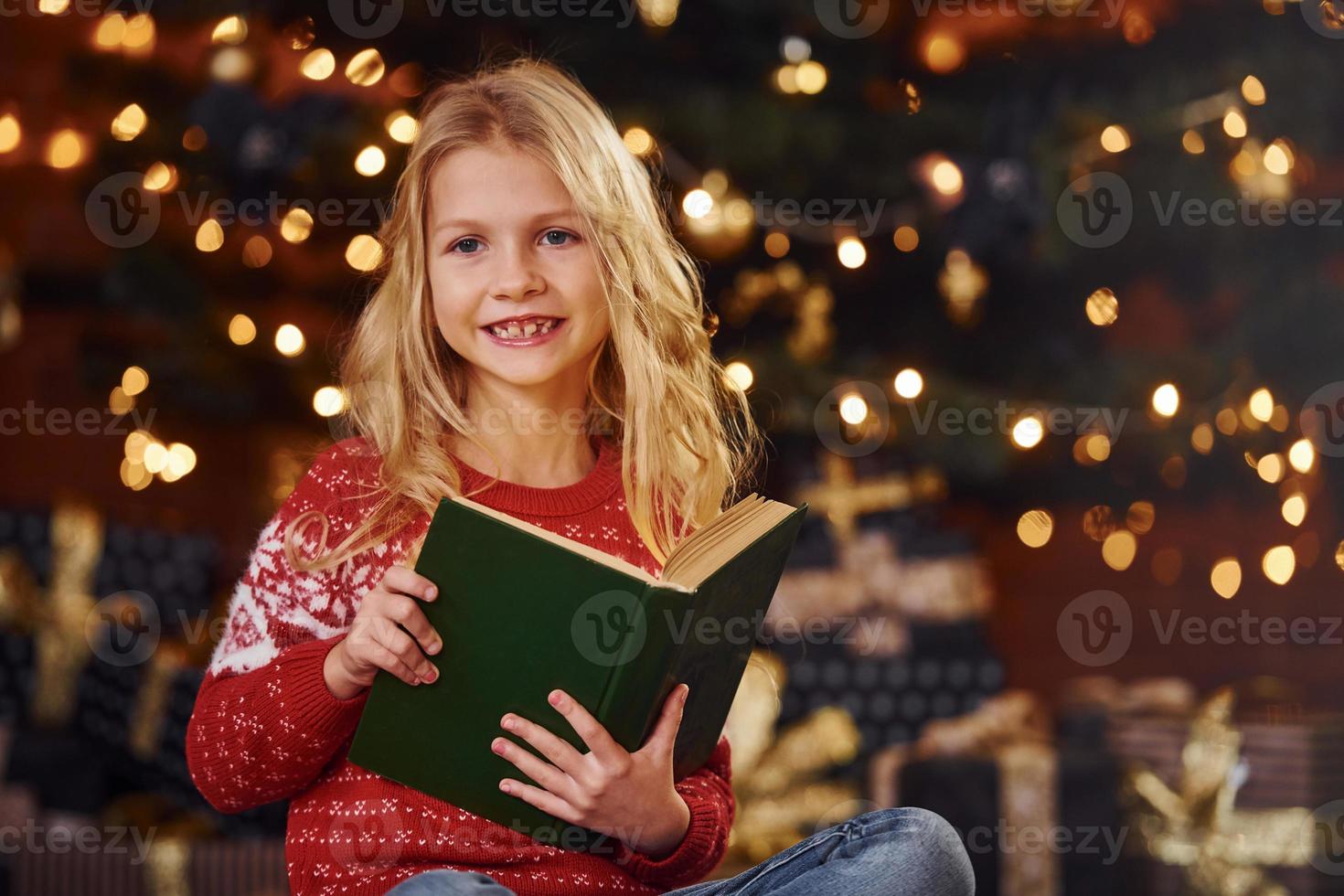 linda niñita con suéter festivo rojo leyendo un libro en el interior en las vacaciones de Navidad foto