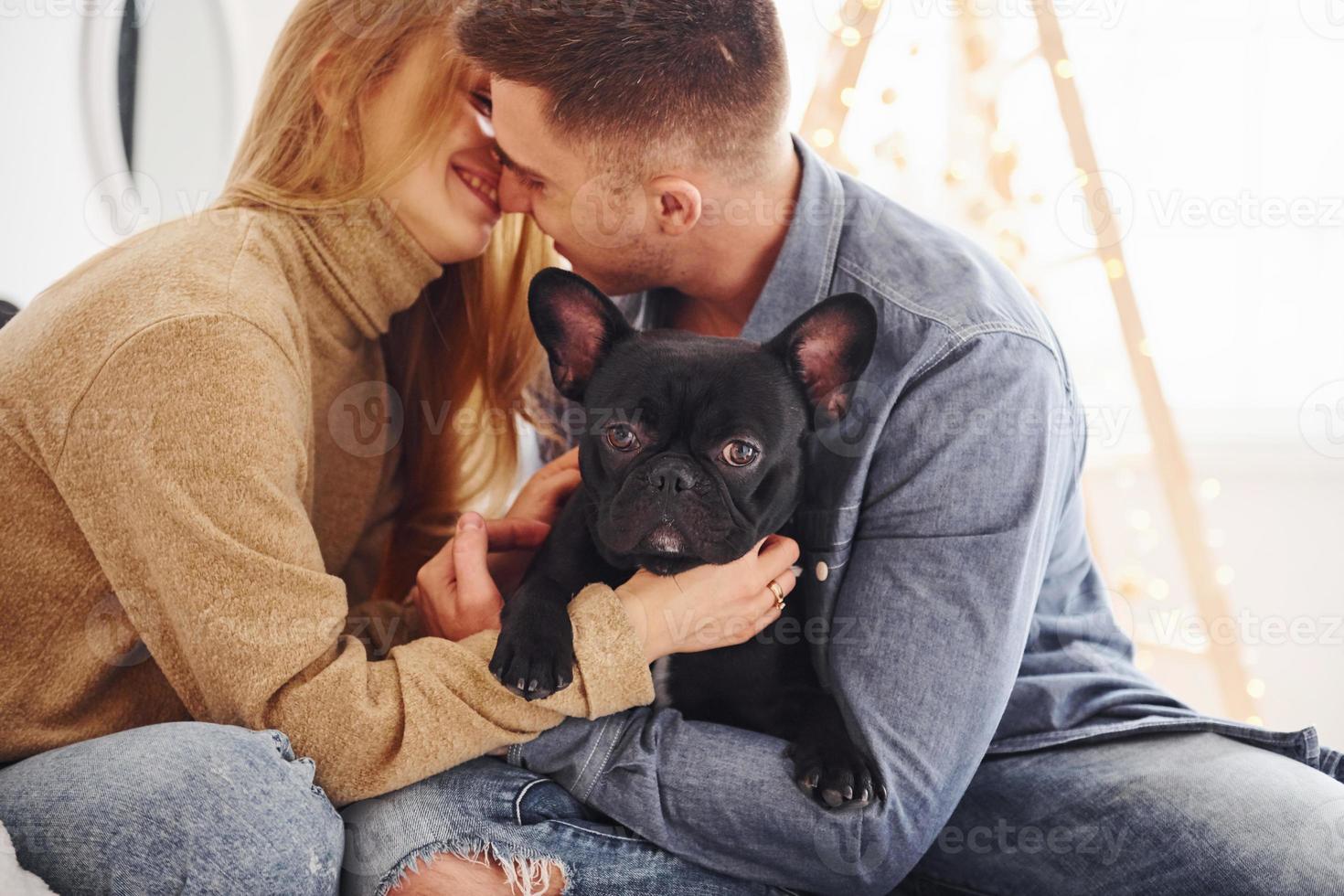 Happy young modern couple sitting on the sofa at home with christmas tree with their cute dog photo