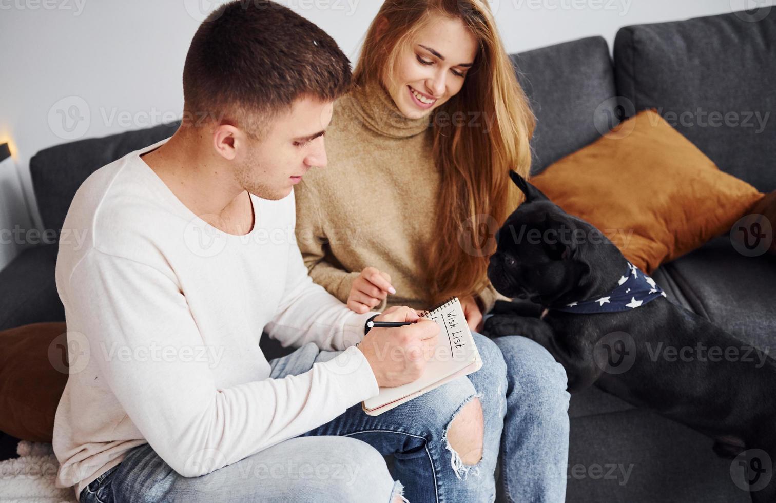 Happy young modern couple sitting on the sofa and writing wishes on notepad at home with their cute dog photo
