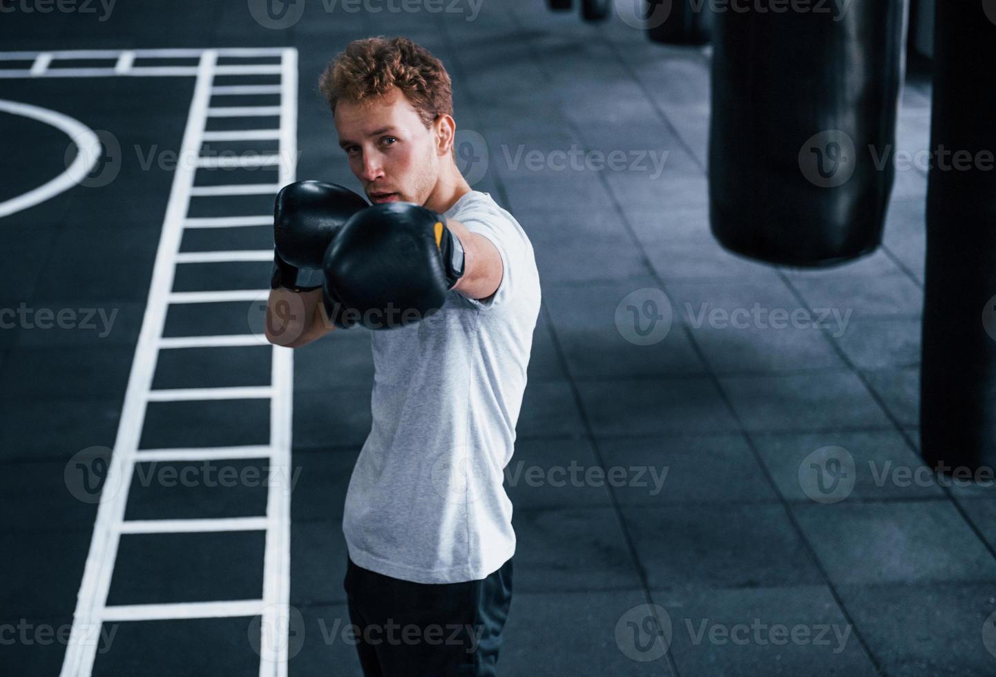 Young man in white shirt and boxing protective gloves doing exercises in gym photo