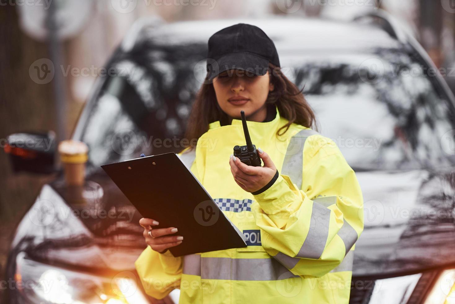 Female police officer in green uniform standing with radio transmitter and notepad against car photo