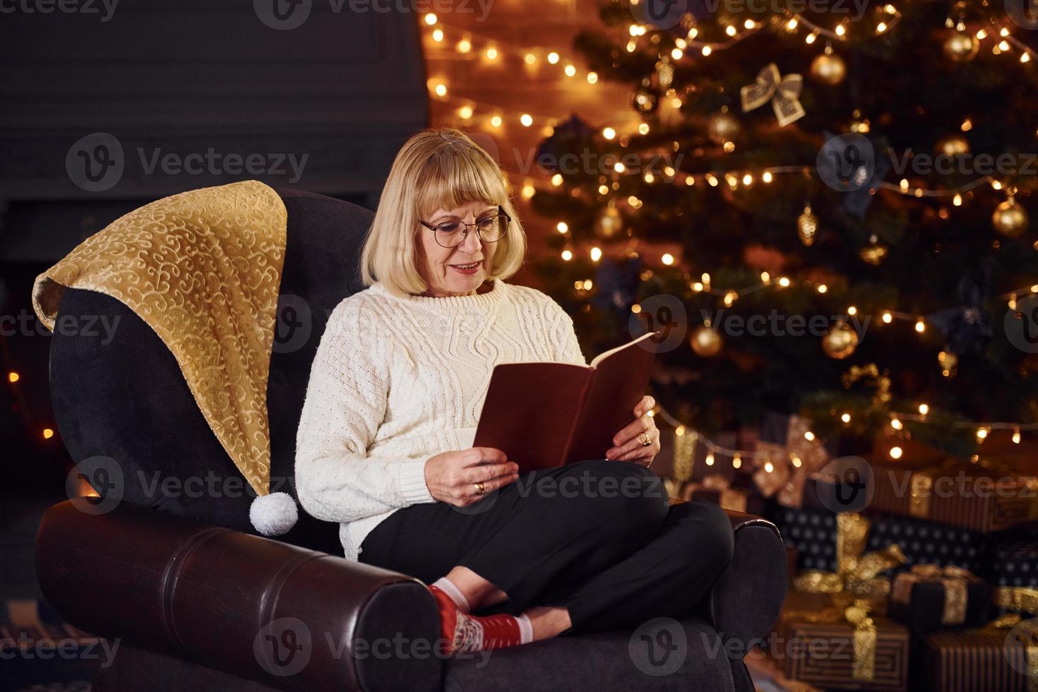 Old woman sitting on the chair with book in festive New year room with christmas decorations photo