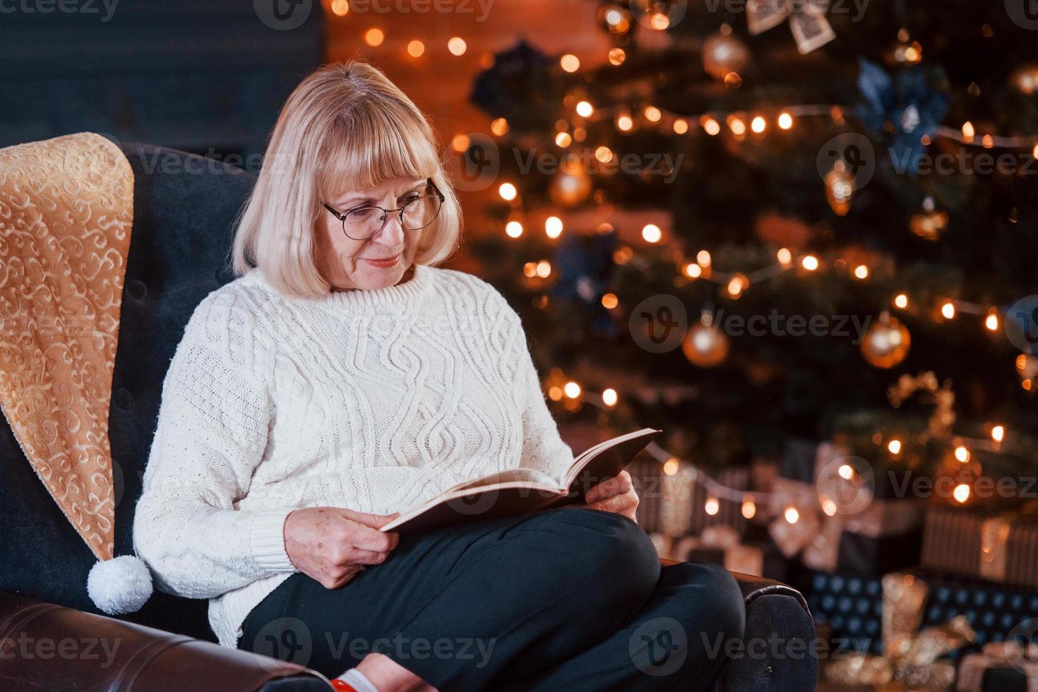 Grandmother sitting on the chair with book in festive New year room with christmas decorations photo