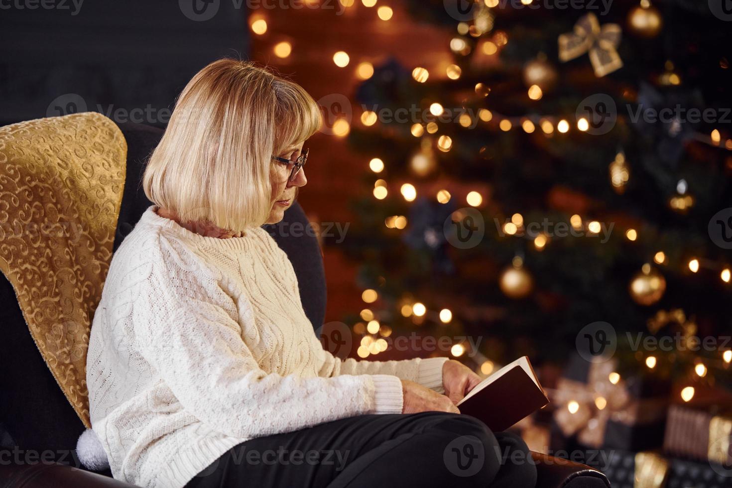 Old woman sitting on the chair with book in festive New year room with christmas decorations photo
