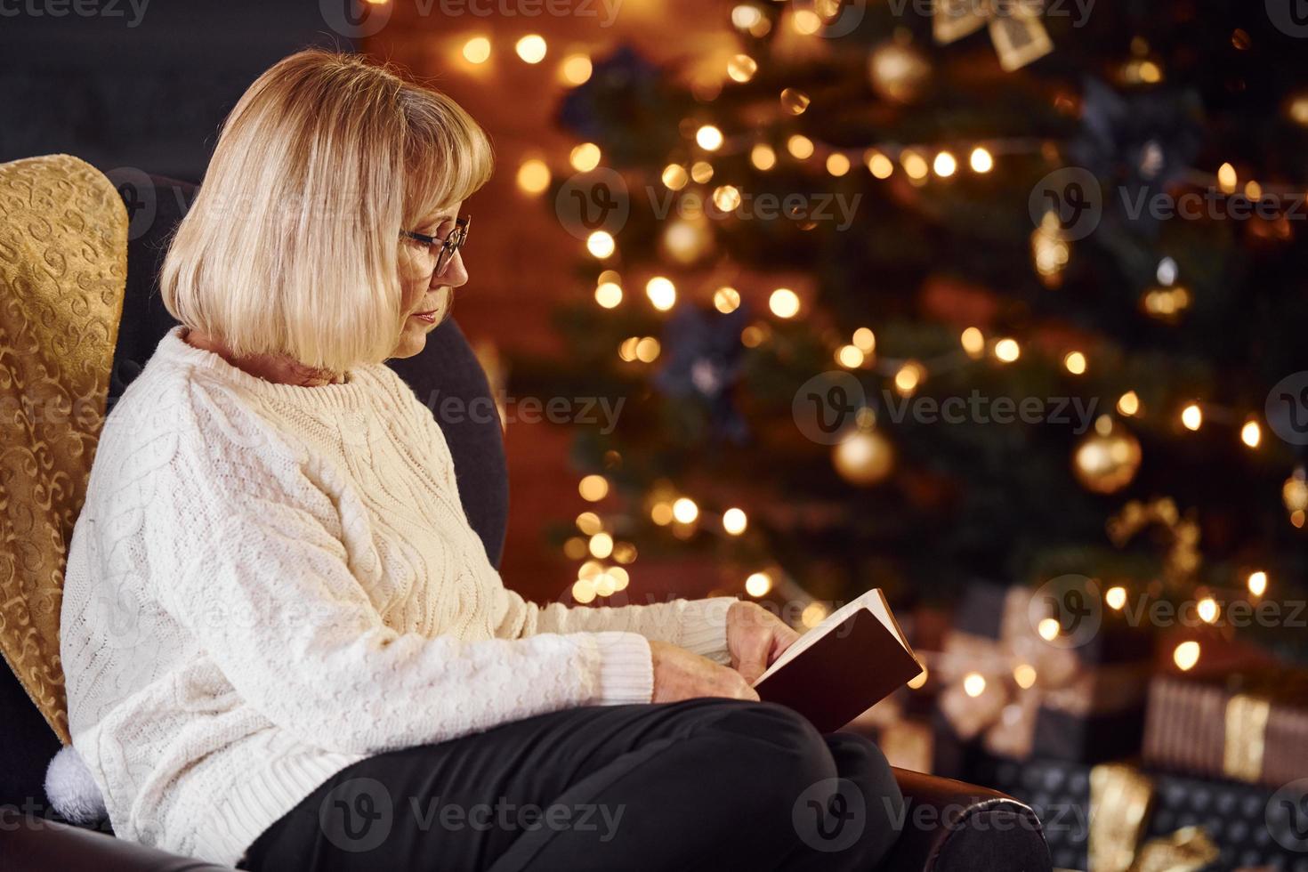Old woman sitting on the chair with book in festive New year room with christmas decorations photo