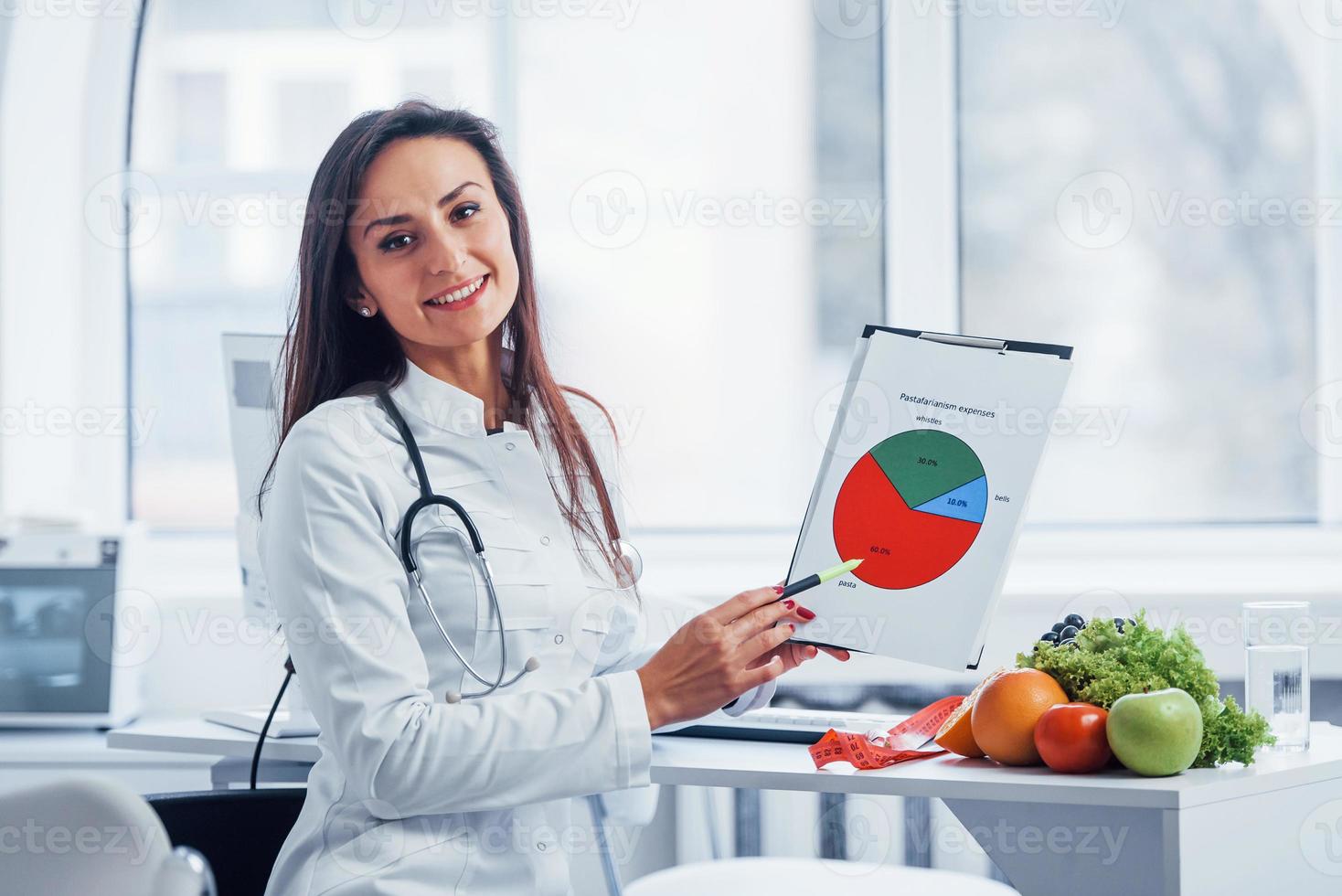 nutricionista femenina con bata blanca sentada en el interior de la oficina en el lugar de trabajo con un gráfico en las manos foto