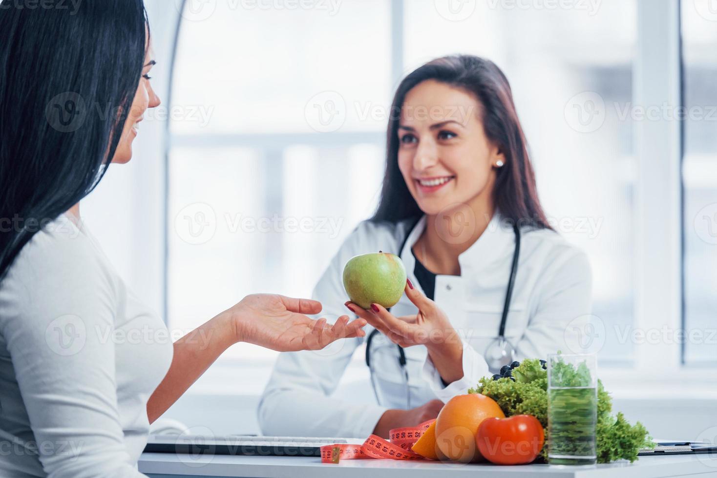 Female nutritionist holding green apple and gives consultation to patient indoors in the office photo