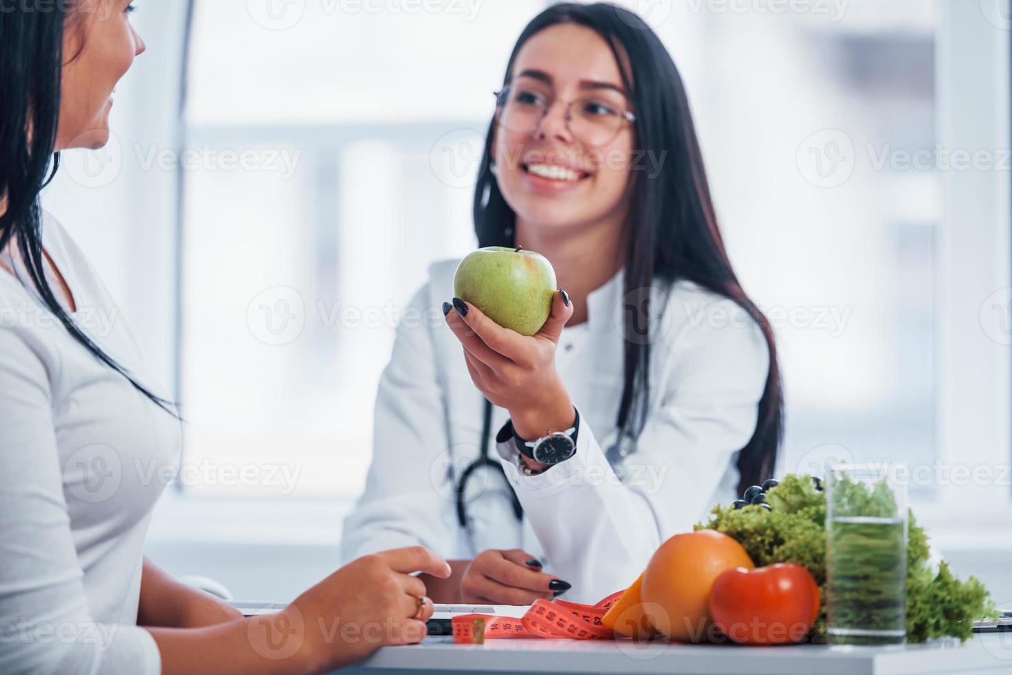 Female nutritionist holding green apple and gives consultation to patient indoors in the office photo