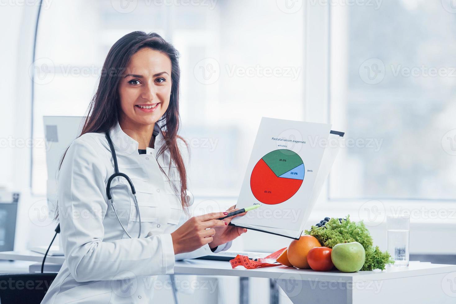 Female nutritionist in white coat sitting indoors in the office at workplace with graph in hands photo