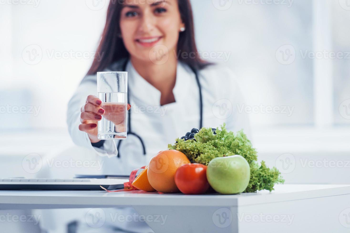 nutricionista femenina con bata blanca sentada en el interior de la oficina en el lugar de trabajo con un vaso de bebida foto