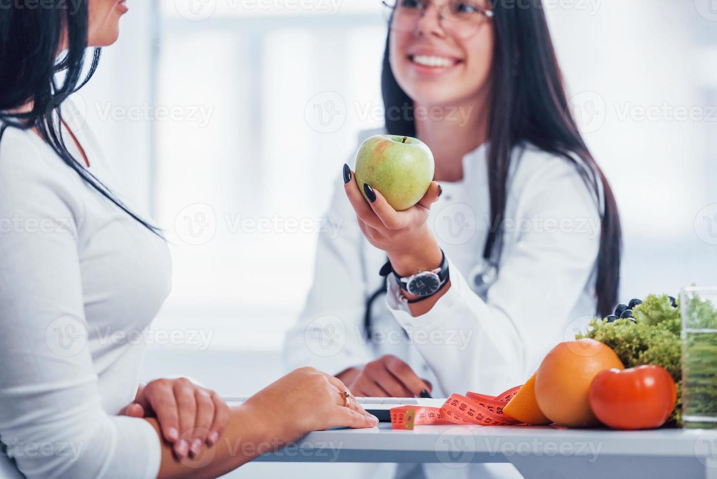 Female nutritionist holding green apple and gives consultation to patient indoors in the office photo