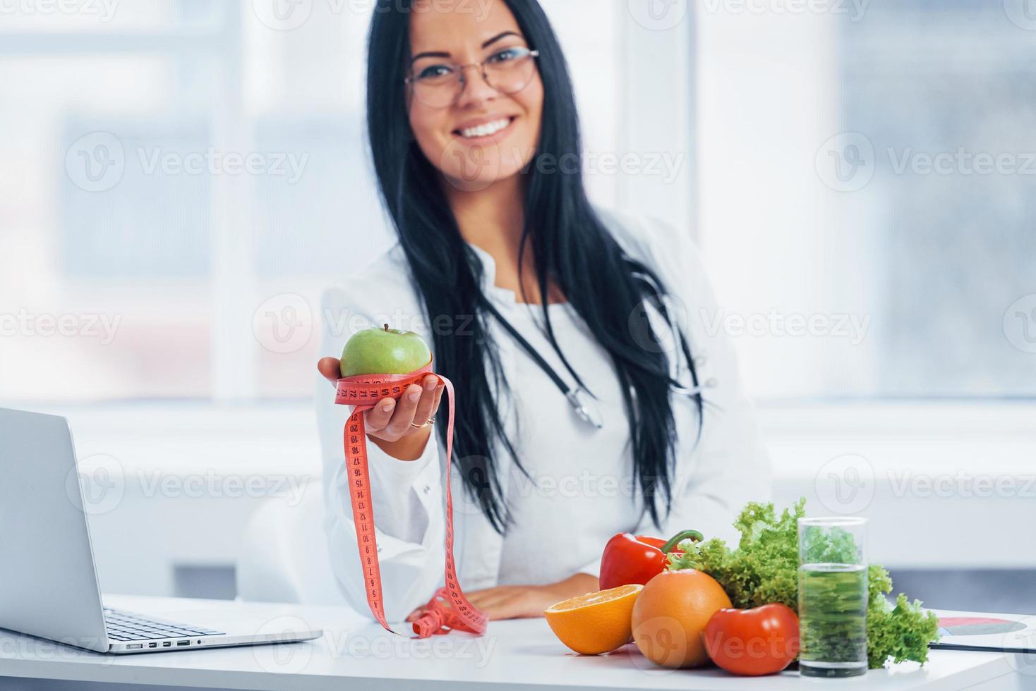 Female nutritionist in white coat holding apple with measuring tape photo