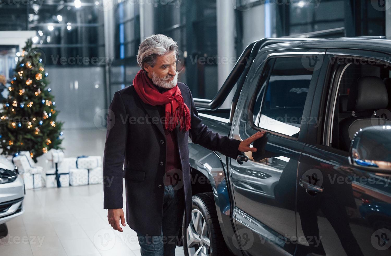 Fashionable old man with grey hair and mustache looking for a new car indoors in salon with christmas tree photo
