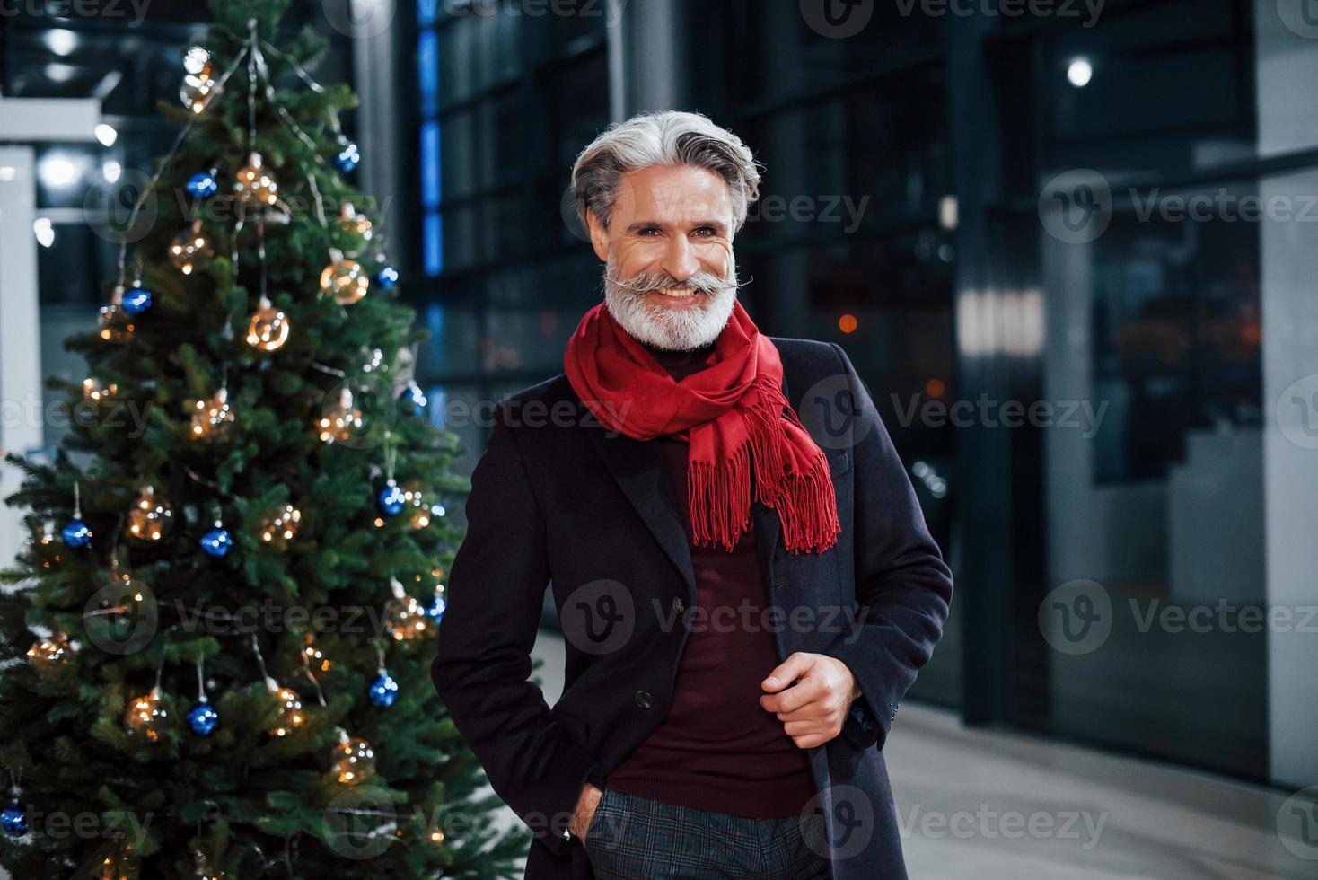 retrato de un anciano de moda con cabello gris y bigote parado cerca de un árbol decorado de navidad foto