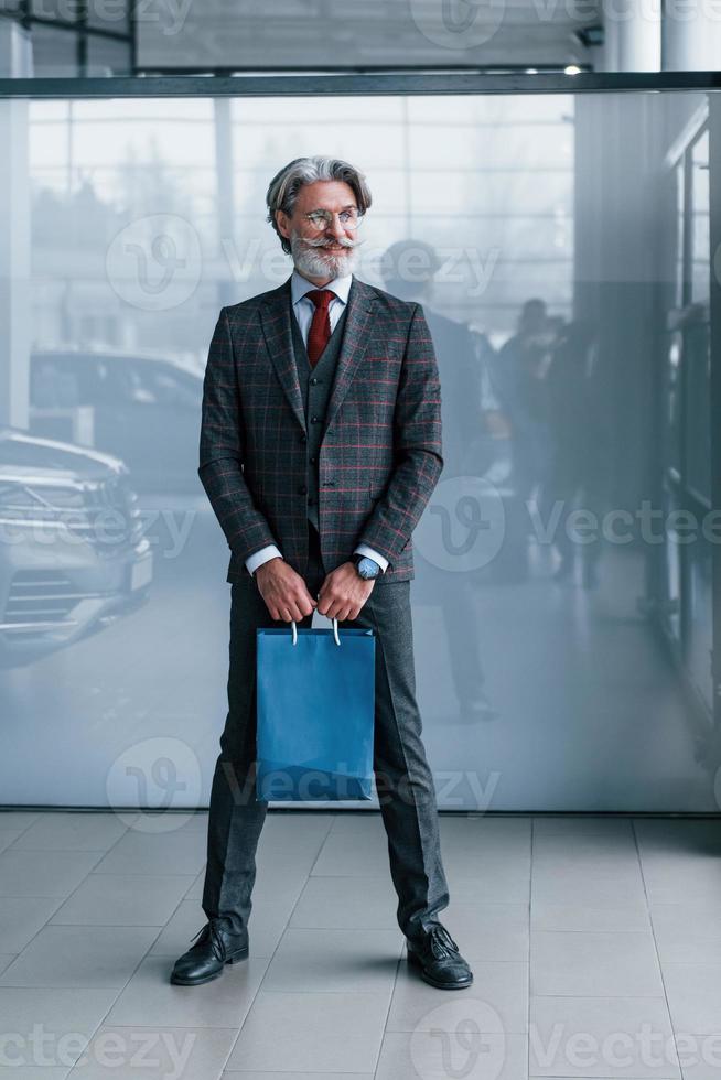 hombre de negocios senior en traje y corbata con pelo gris y barba de pie en el interior con paquete azul foto