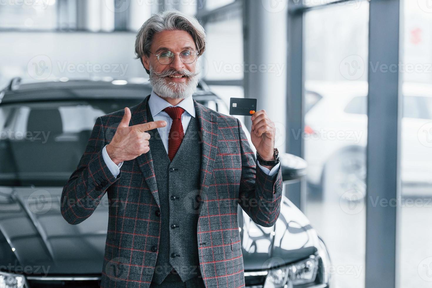 Man with grey hair and mustache standing against modern car indoors with credit card in hand photo