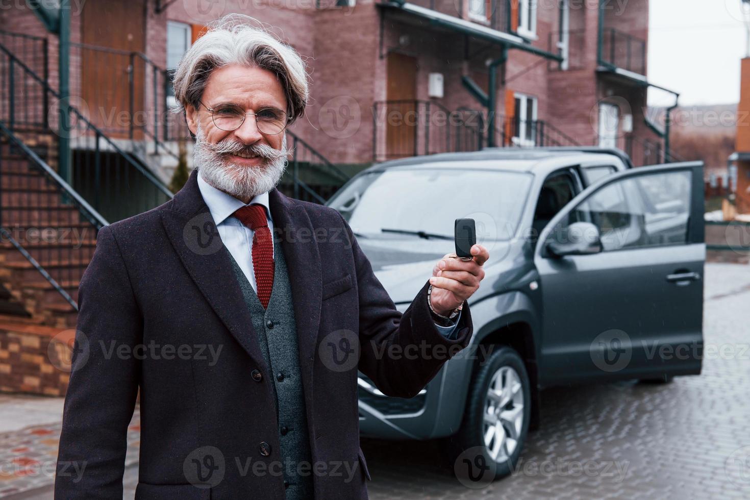 Fashionable senior man with gray hair and beard standing outdoors on the street near his car with keys in hand photo
