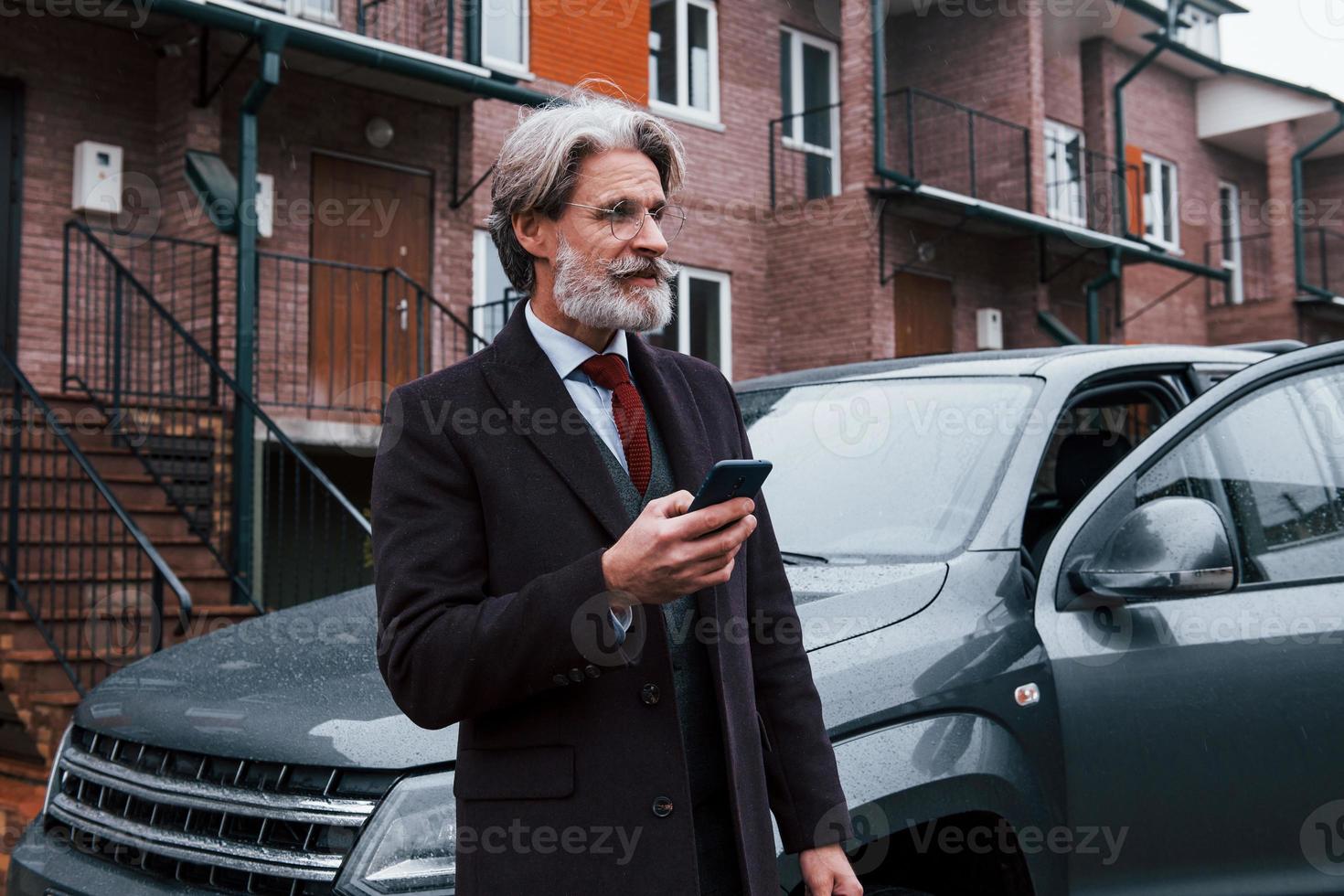 Fashionable senior man with gray hair and beard standing with phone near his car on the street photo