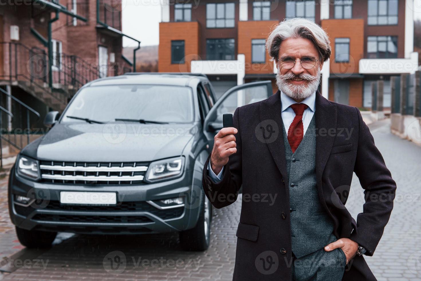 Fashionable senior man with gray hair and beard standing outdoors on the street near his car with keys in hand photo