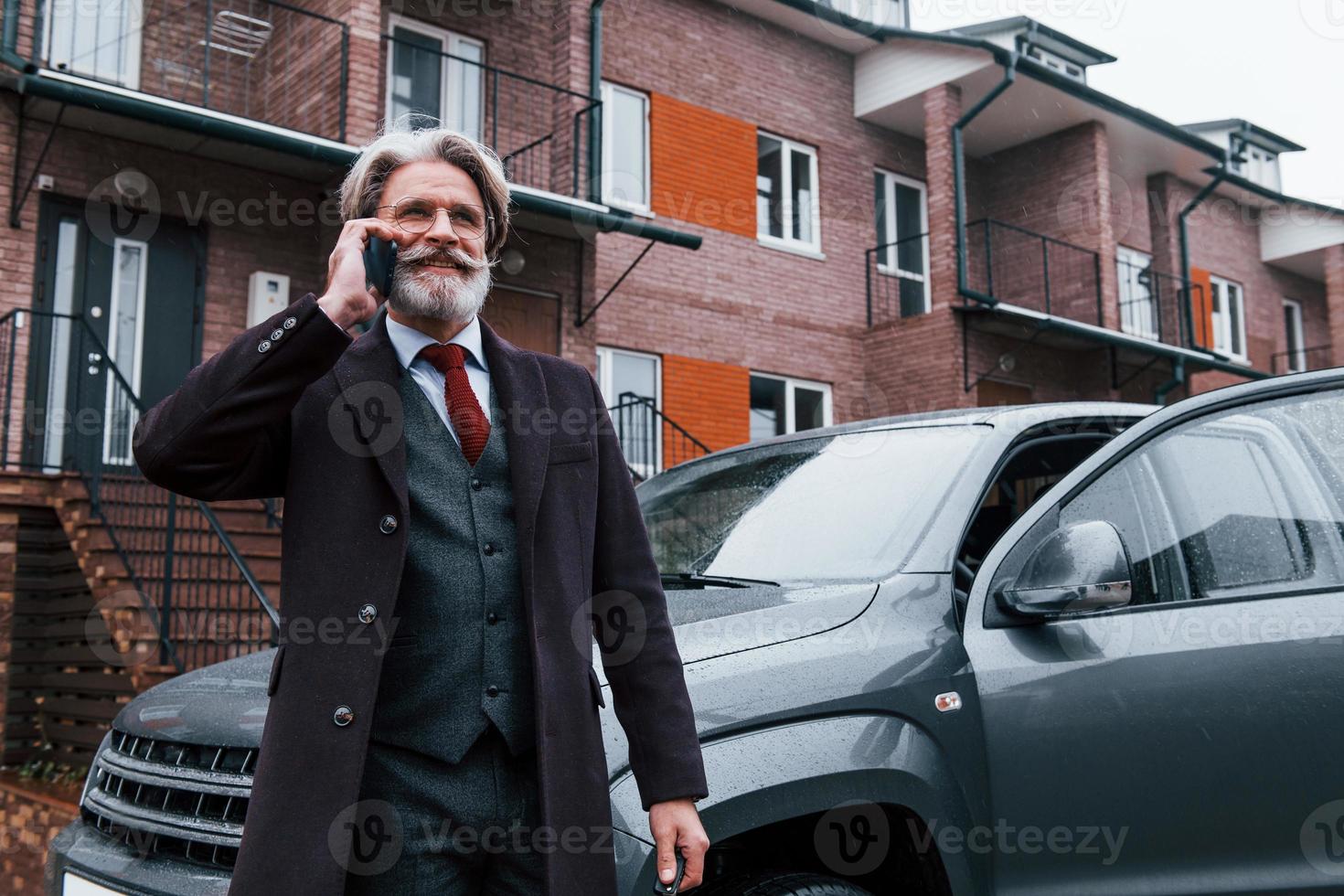 Fashionable senior man with gray hair and beard have conversation by phone outdoors on the street near his car photo