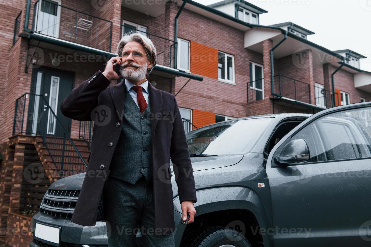 un anciano de moda con cabello gris y barba tiene una conversación por teléfono al aire libre en la calle cerca de su auto foto