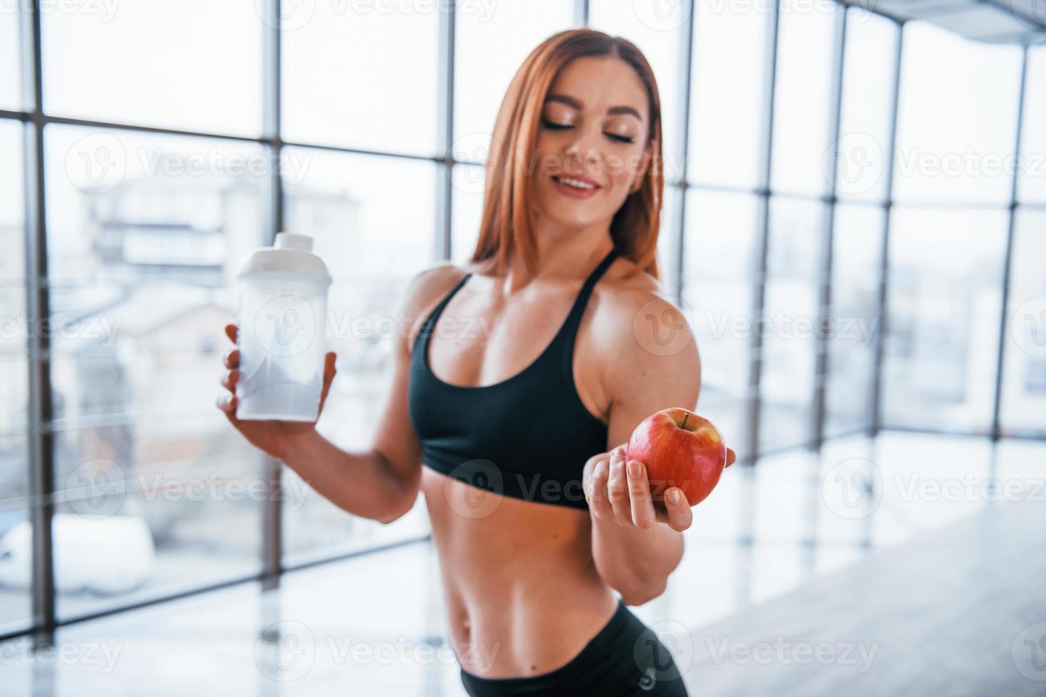 Sportive woman with nice body stands indoors with bottle of water and fresh apple in hands photo