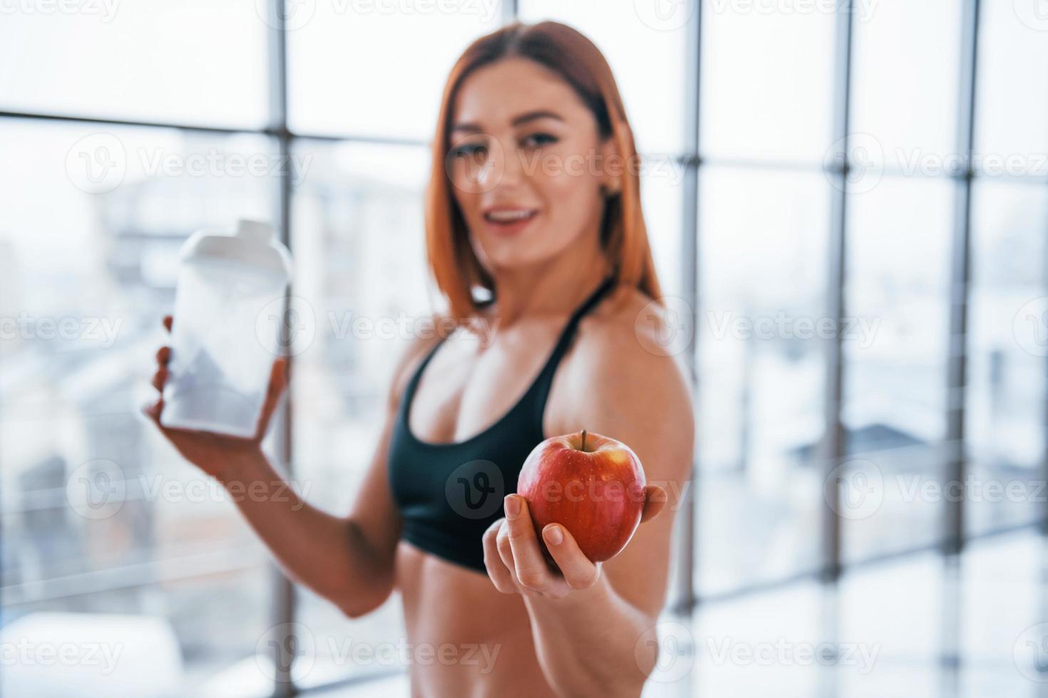 Sportive woman with nice body stands indoors with bottle of water and fresh apple in hands photo