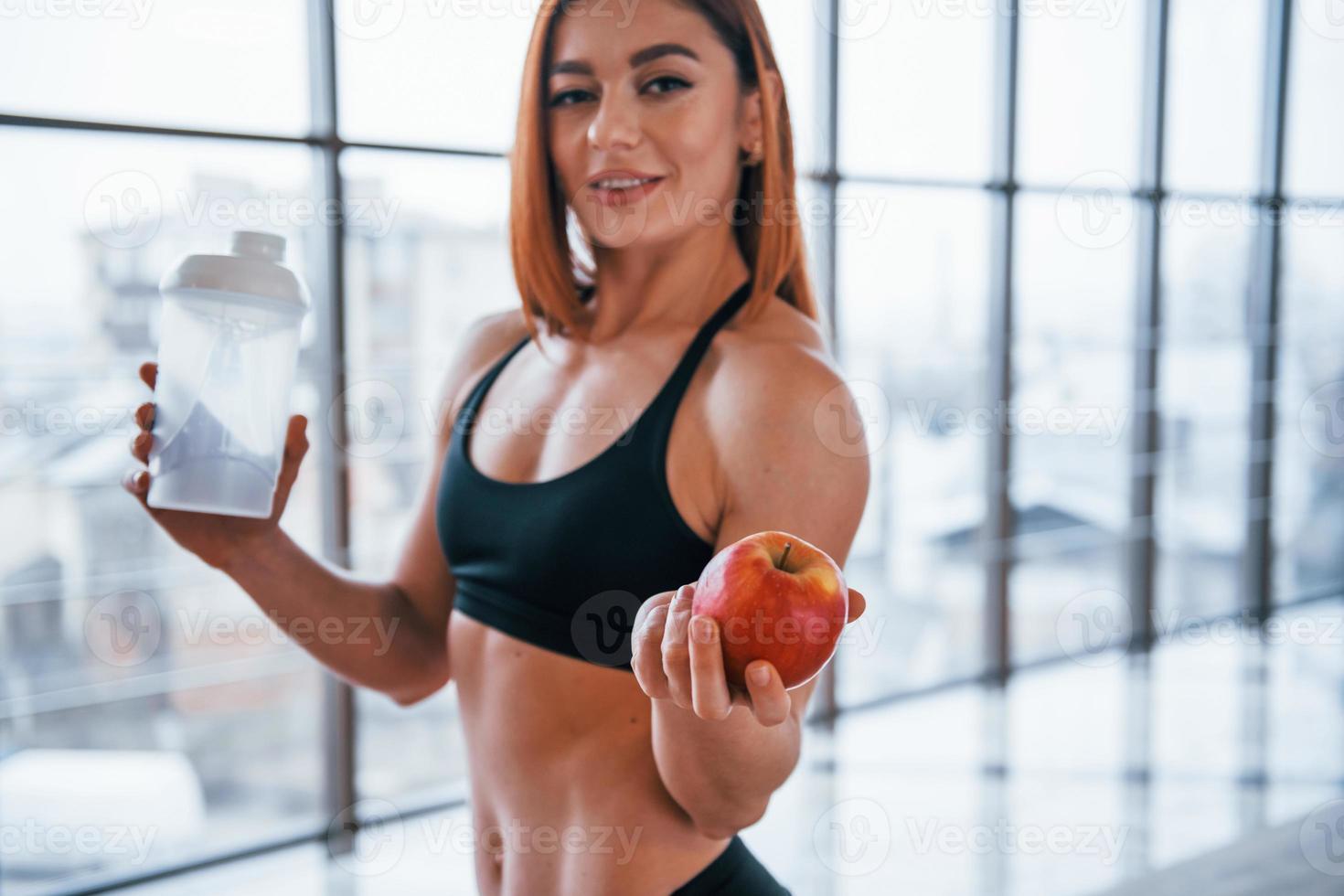 Sportive woman with nice body stands indoors with bottle of water and fresh apple in hands photo