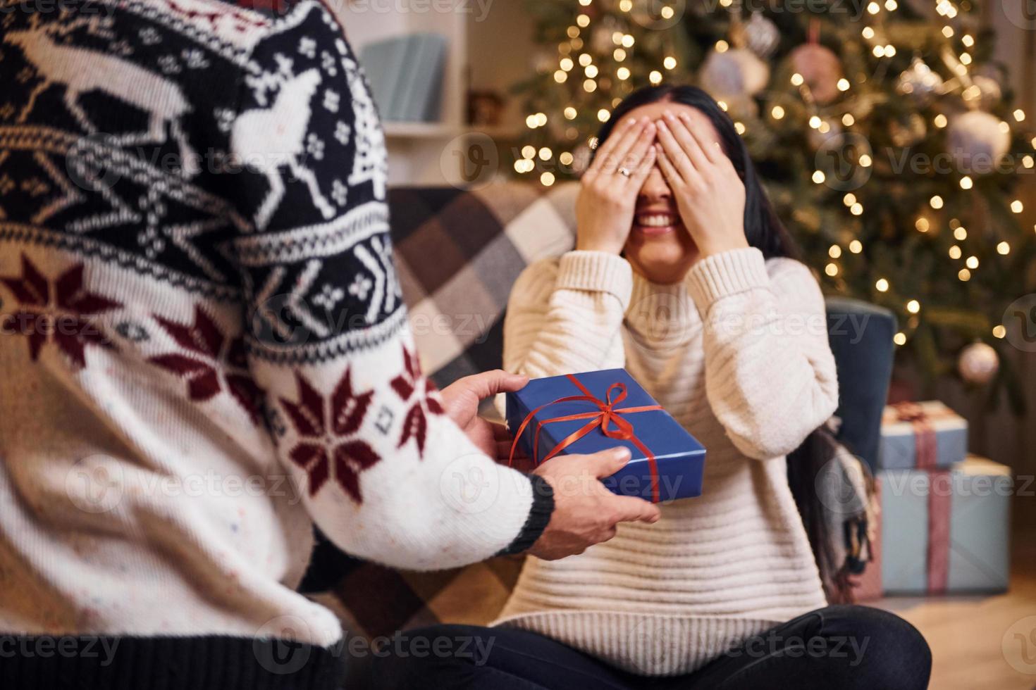 el hombre cubre los ojos de su novia y le da una sorpresa de año nuevo en una habitación decorada con navidad foto