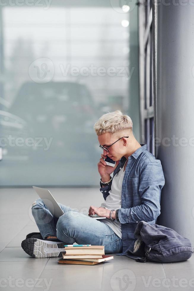 Young hipster guy with phone sitting indoors near grey background with laptop and books photo