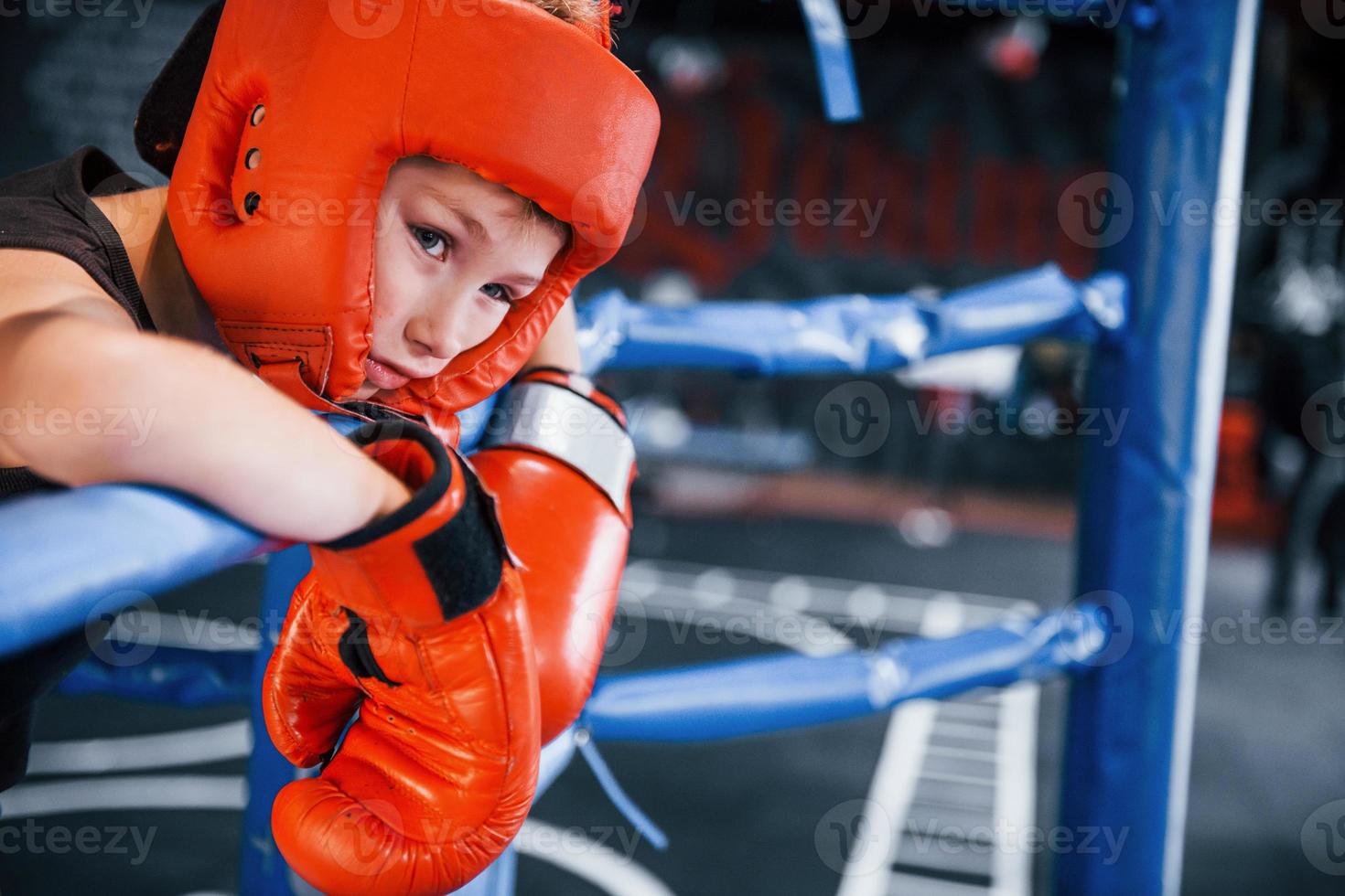 niño cansado en equipo de protección apoyado en los nudos del ring de boxeo foto