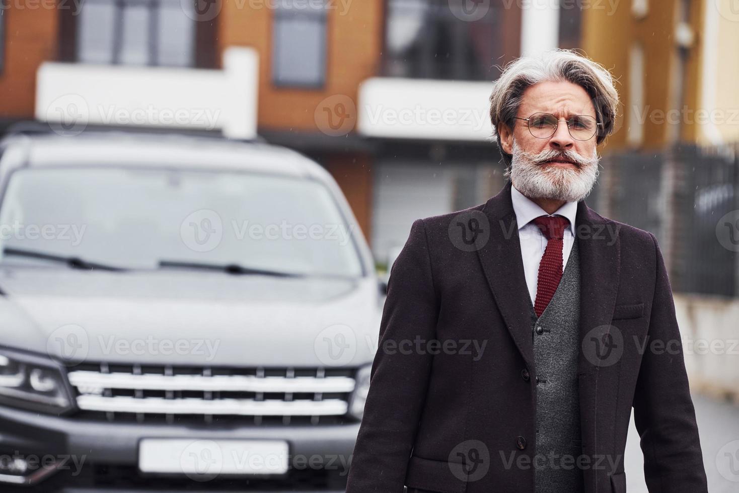 Fashionable senior man with gray hair and beard walking outdoors on the street near his car photo