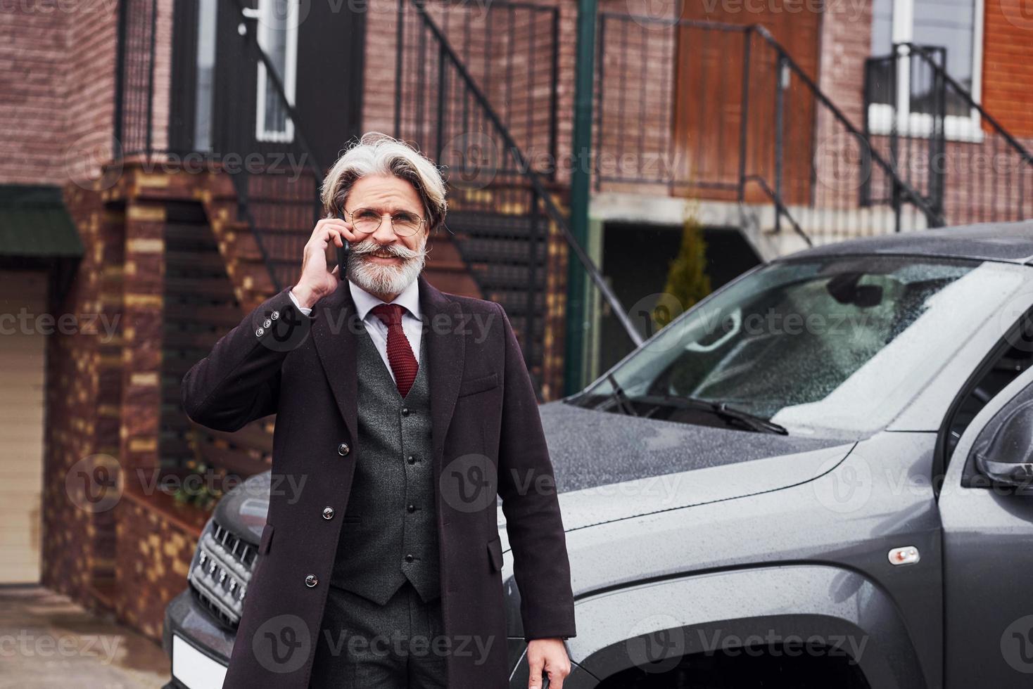 Fashionable senior man with gray hair and beard is outdoors on the street near his car have conversation by the phone photo