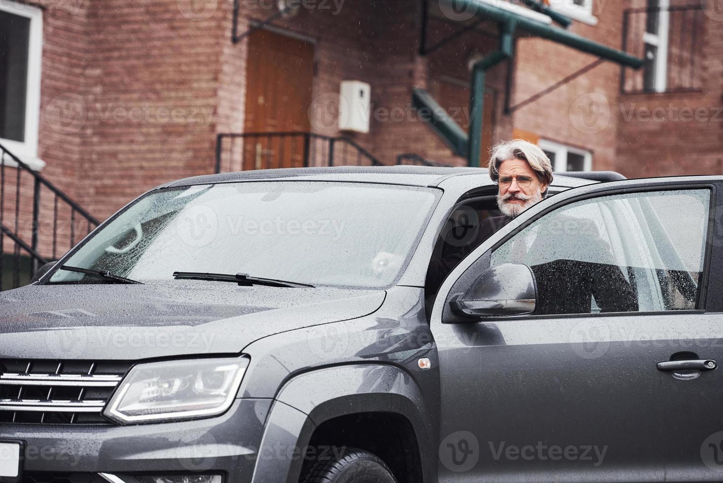 un anciano de moda con pelo gris y barba está al aire libre en la calle sentado en su coche foto