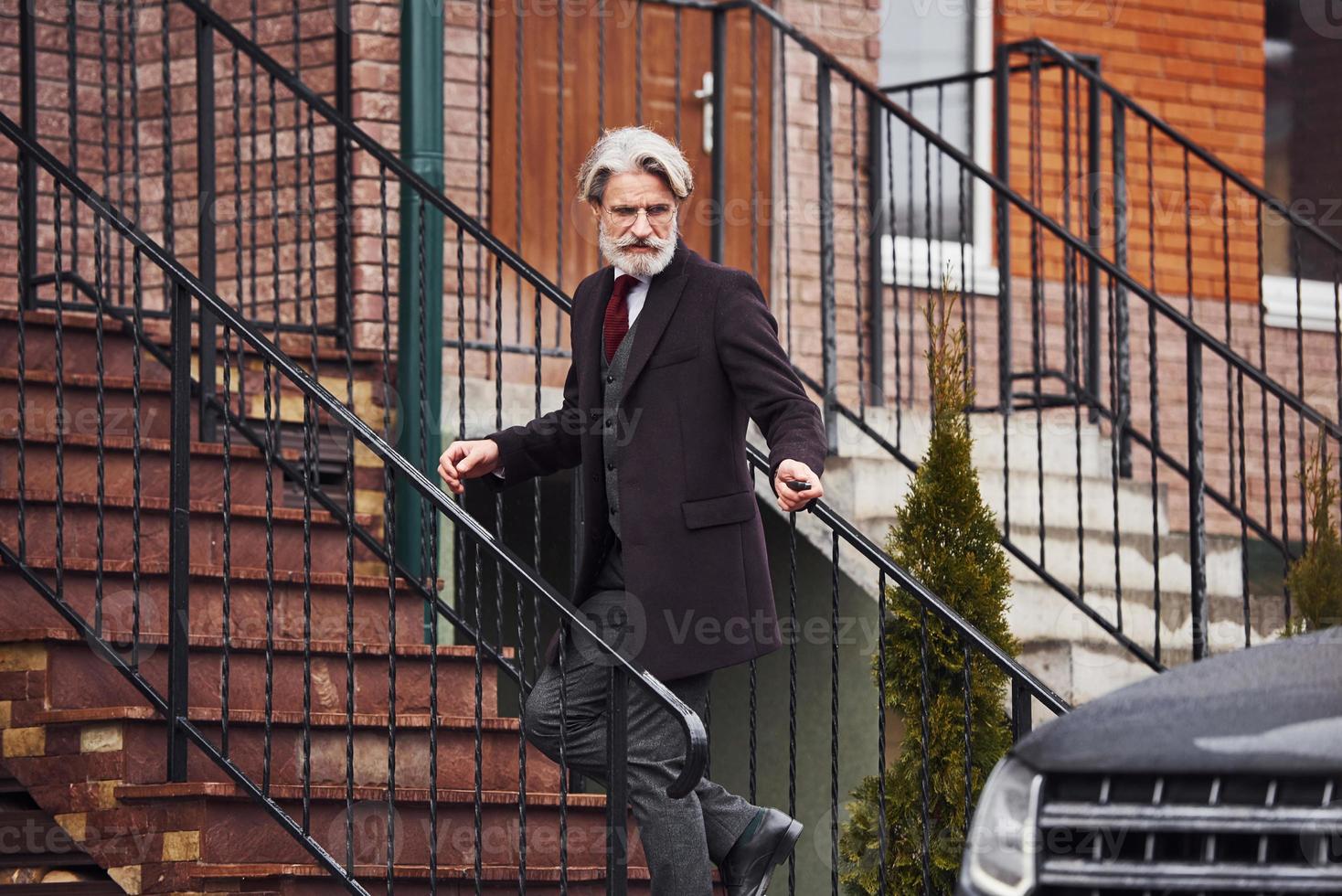 Fashionable senior man with gray hair and beard is outdoors on the street near his car photo