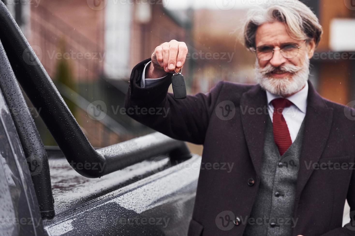 hombre mayor de moda con pelo gris y barba parado afuera en la calle cerca de su auto con llaves en la mano foto