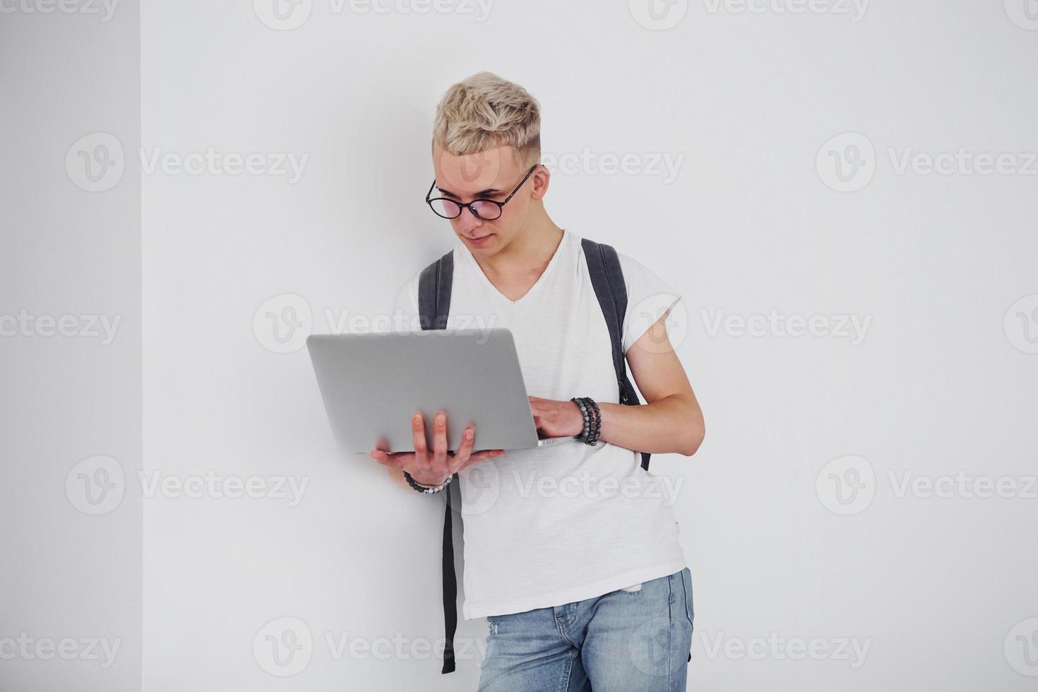 Student in casual clothes and with backpack stands indoors against white wall with laptop photo