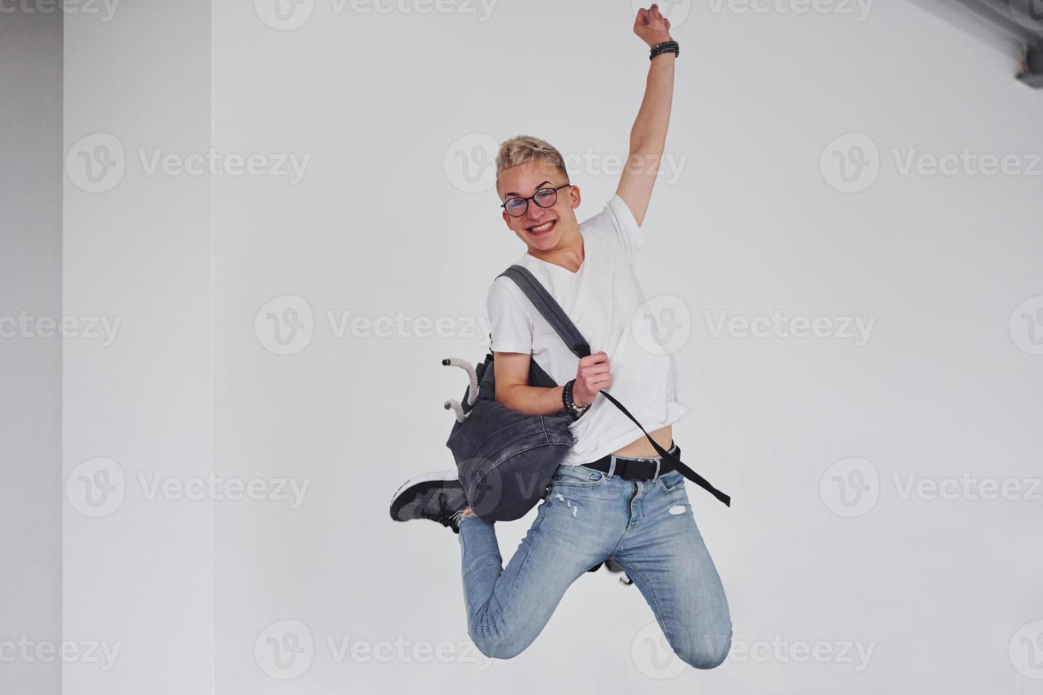 Happy hipster guy in casual clothes and with backpack jumping indoors against white wall photo