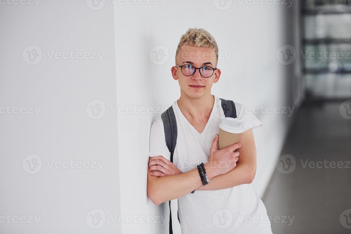 Student in casual clothes and with backpack stands indoors against white wall with cup of drink photo