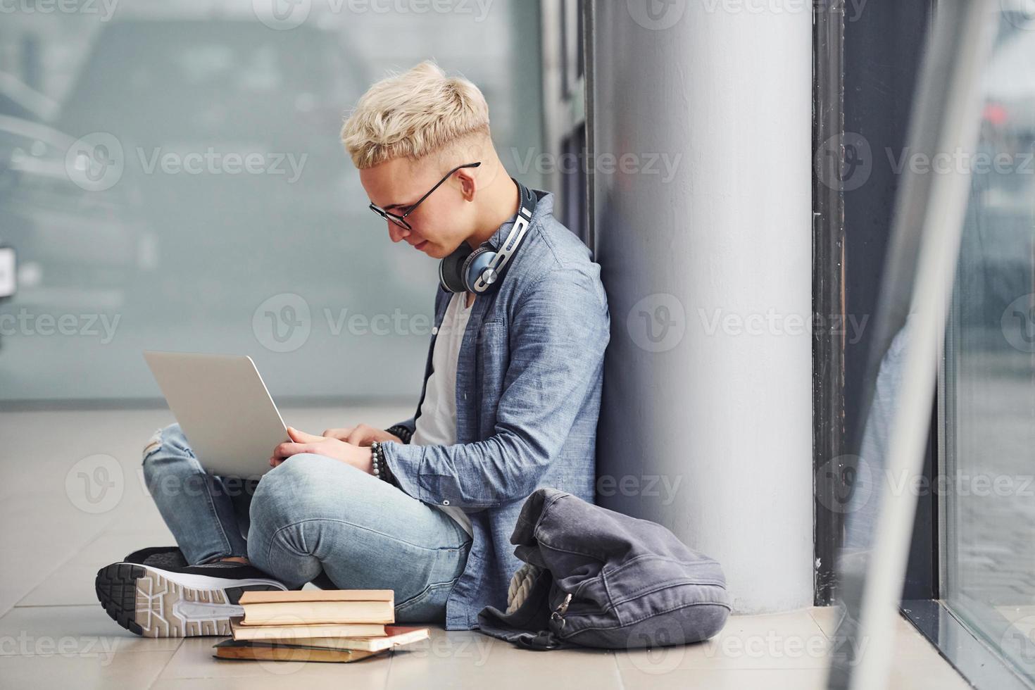 Young hipster guy sitting indoors near grey background with laptop and books photo