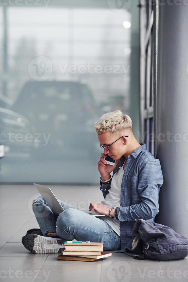 Young hipster guy with phone sitting indoors near grey background with laptop and books photo
