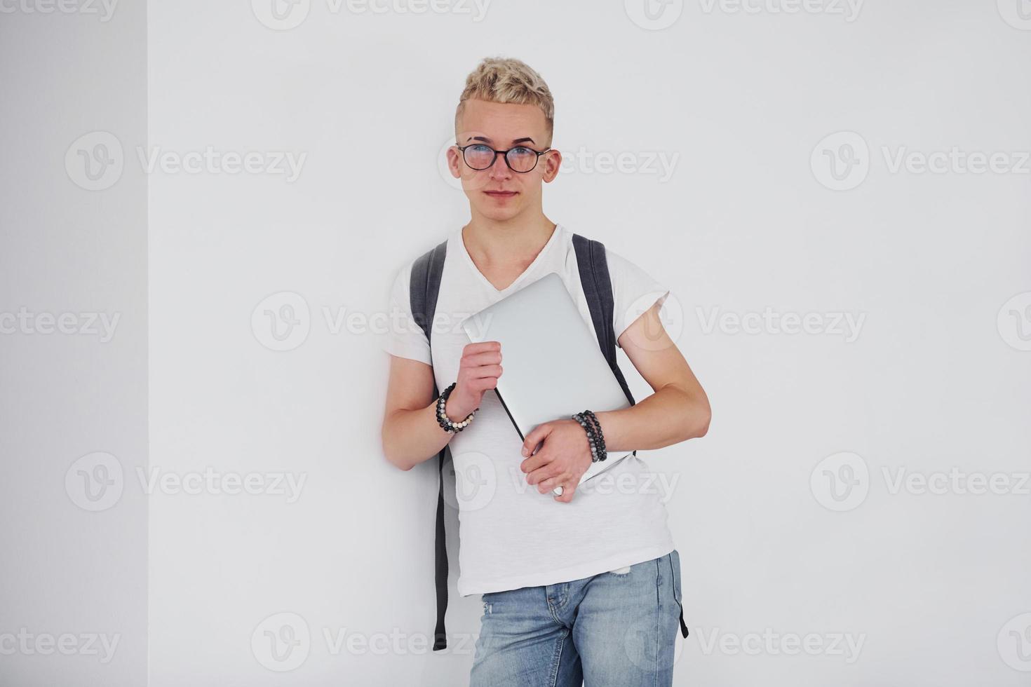 Student in casual clothes and with backpack stands indoors against white wall with laptop photo