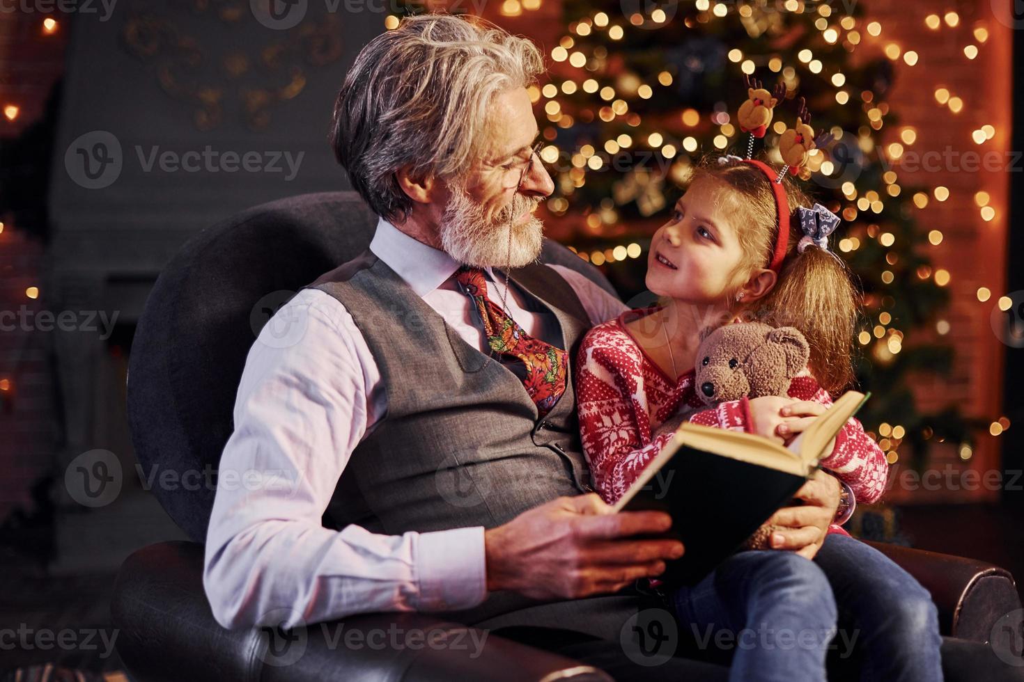 un anciano alegre con pelo gris y barba sentado con una niña en una sala de Navidad decorada leyendo un libro foto