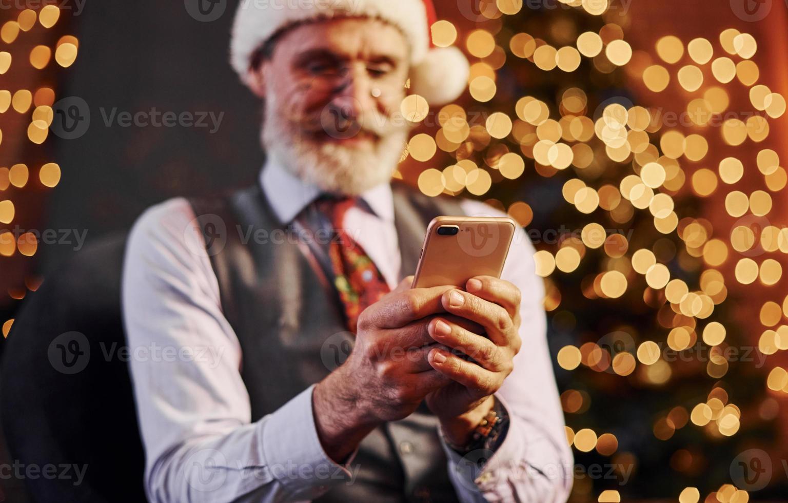 Portrait of stylish senior with grey hair and beard sitting in decorated room and in christmas hat photo