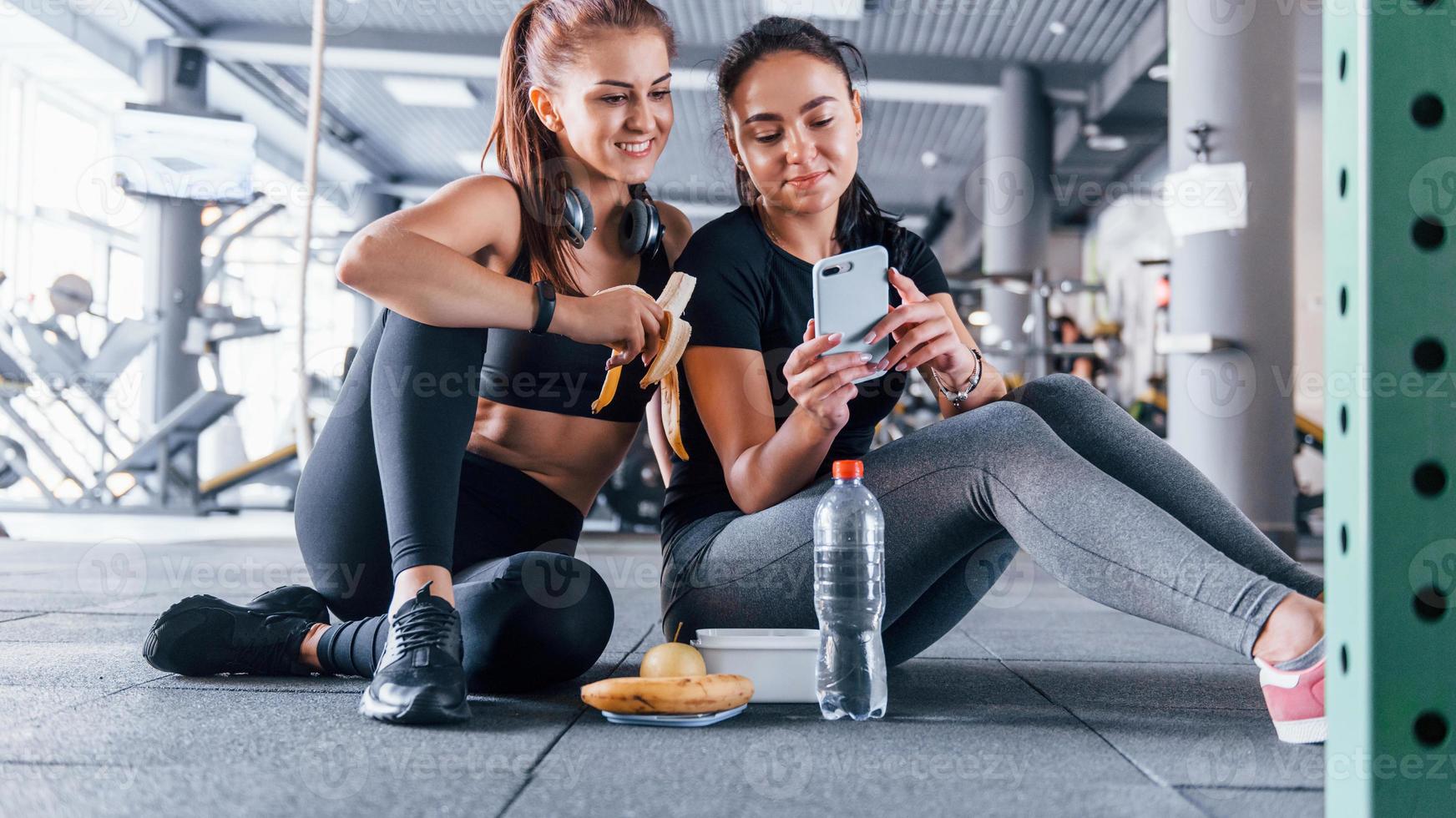 dos amigas con ropa deportiva están en el gimnasio comiendo frutas y usando el teléfono foto