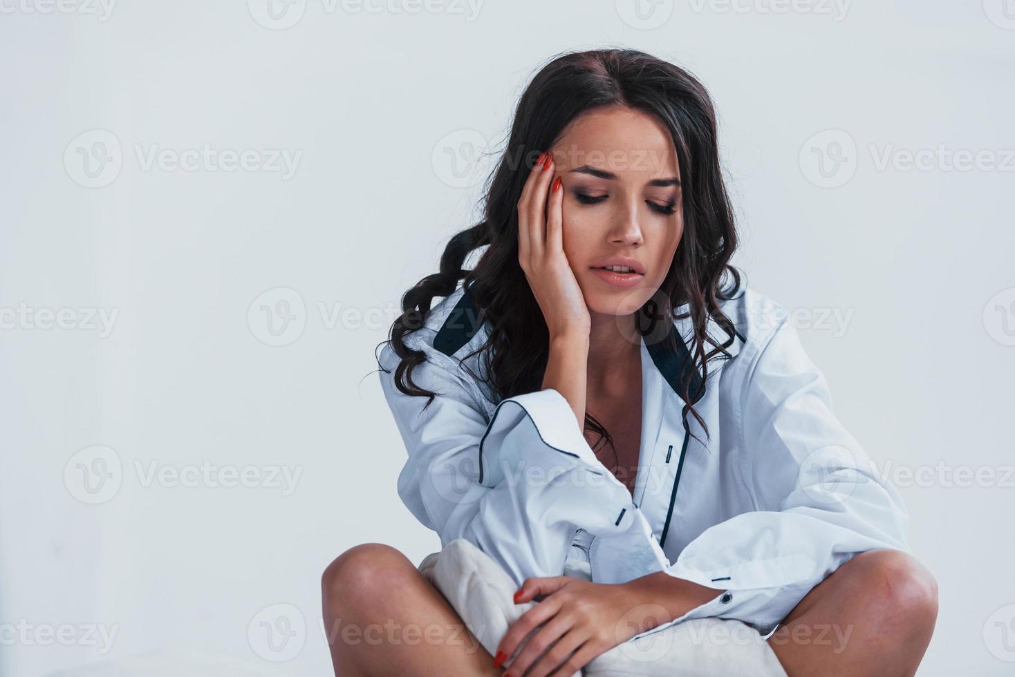 Tired and stressful brunette in white coat sitting on the bed in white room photo