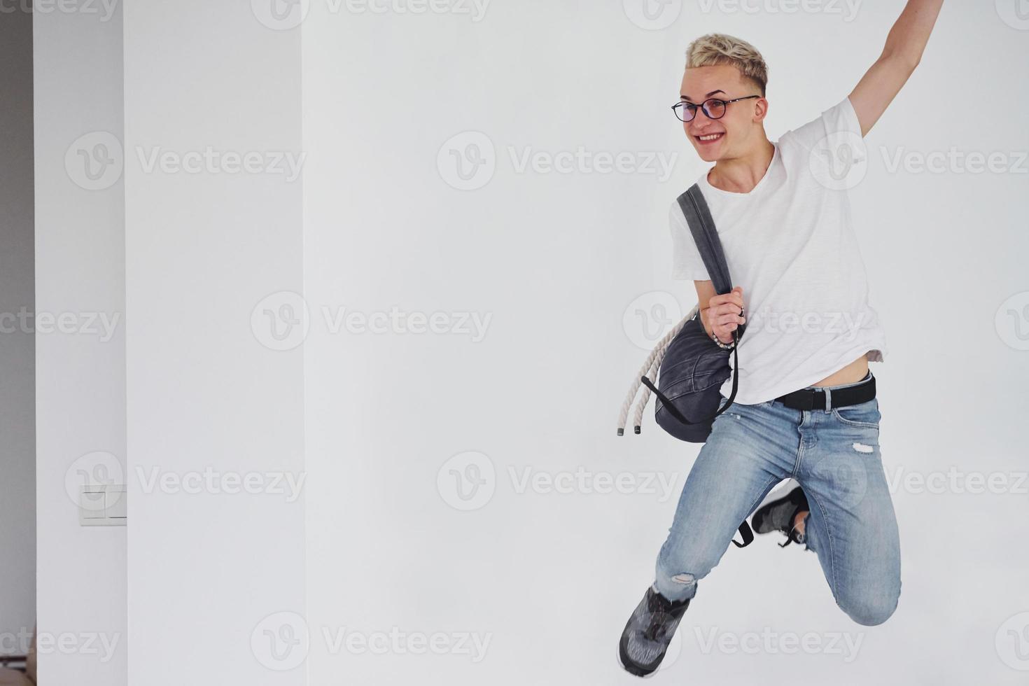 Happy hipster guy in casual clothes and with backpack jumping indoors against white wall photo