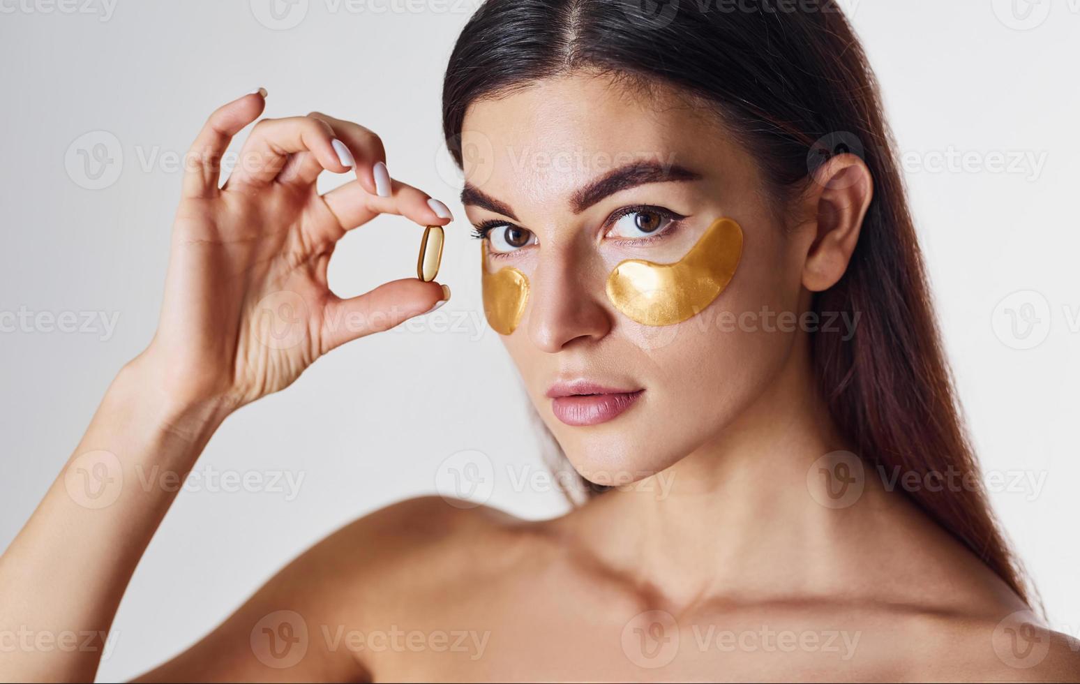 mujer desnuda en el estudio con cintas doradas cerca de los ojos y con una pastilla en la mano foto